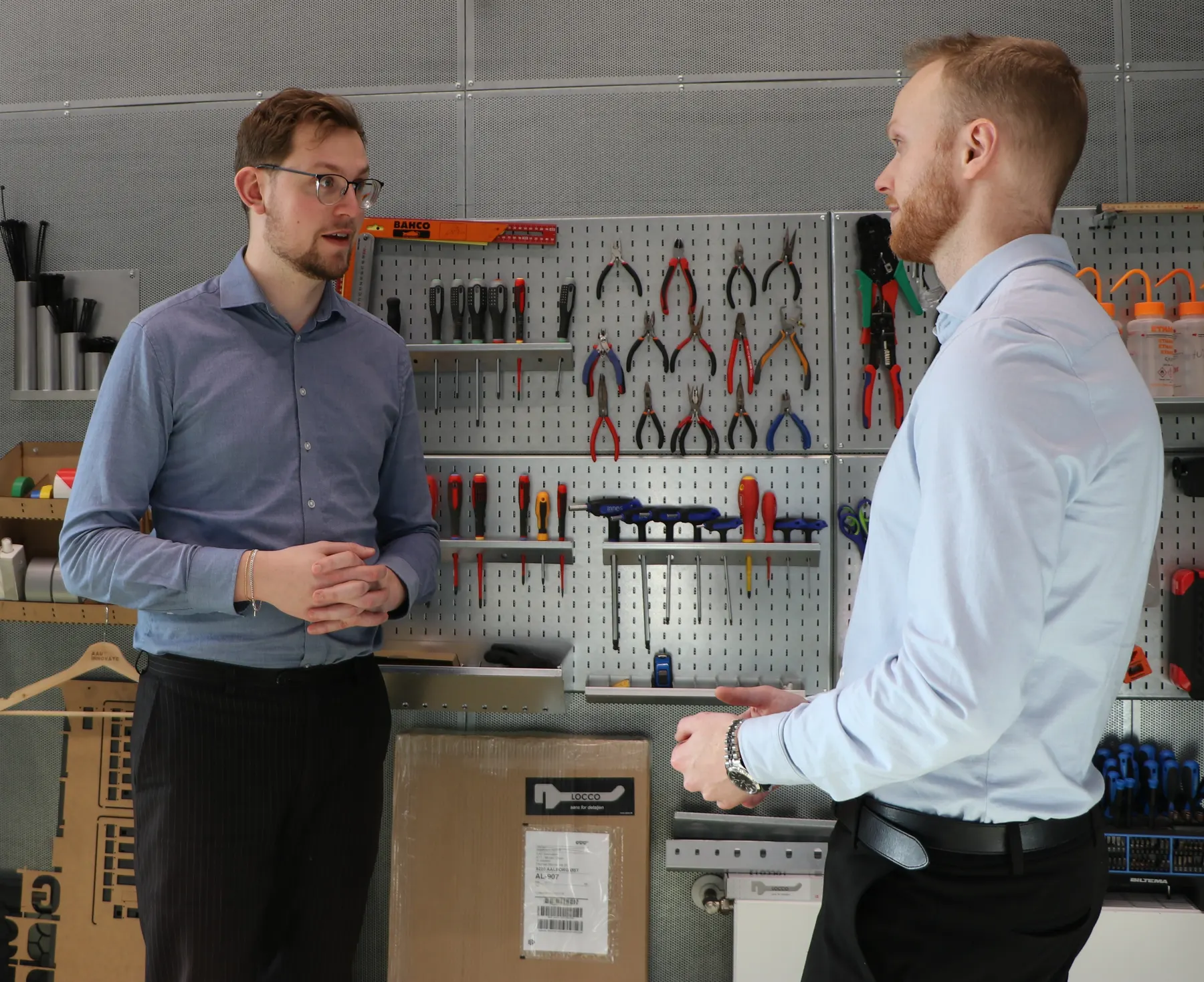Two people talking in front of a wall of tools in a workshop